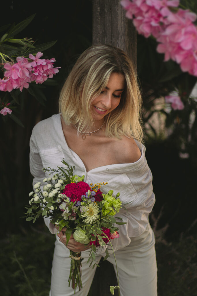 femme en blanc tenant un bouquet de fleurs appuyée sur un poteau avec des laurier rose en fond